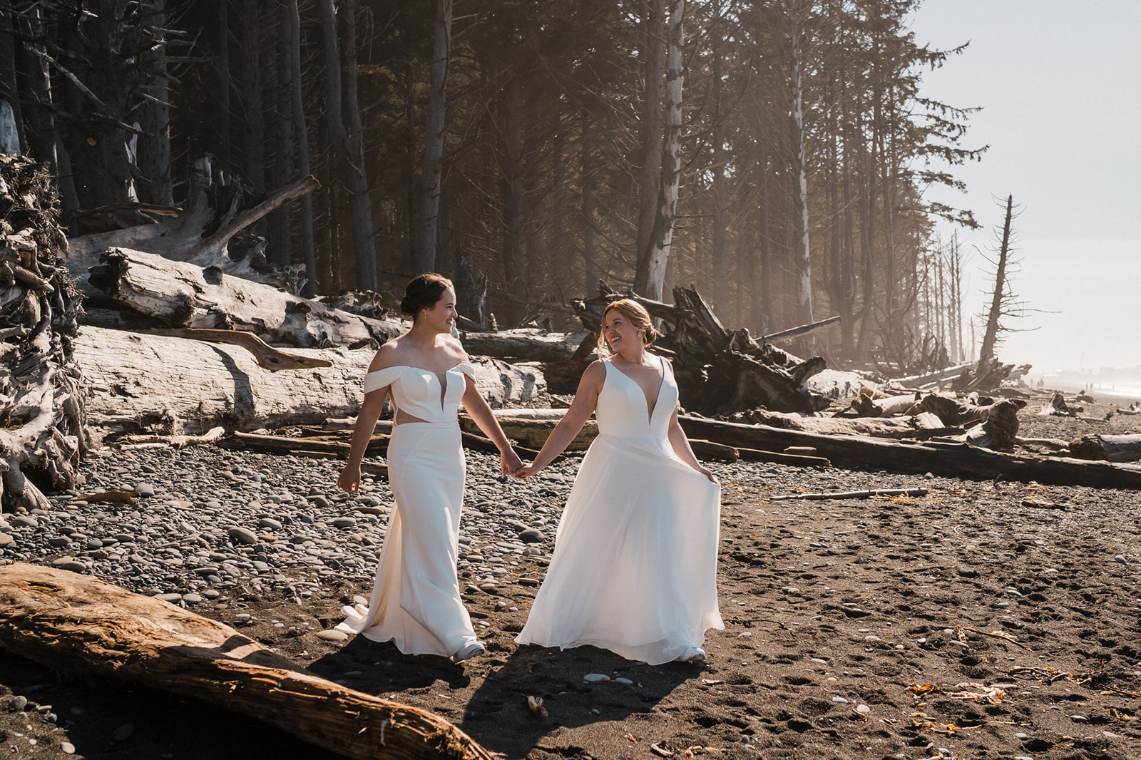 Two brides hold hands while walking across the beach in their white elopement dresses. 