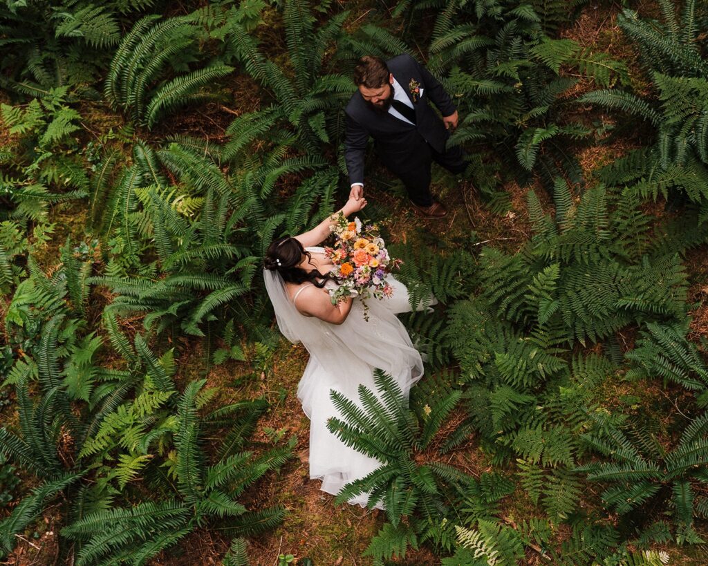 Bride and groom hold hands while walking through a trail filled with ferns in the forest. 