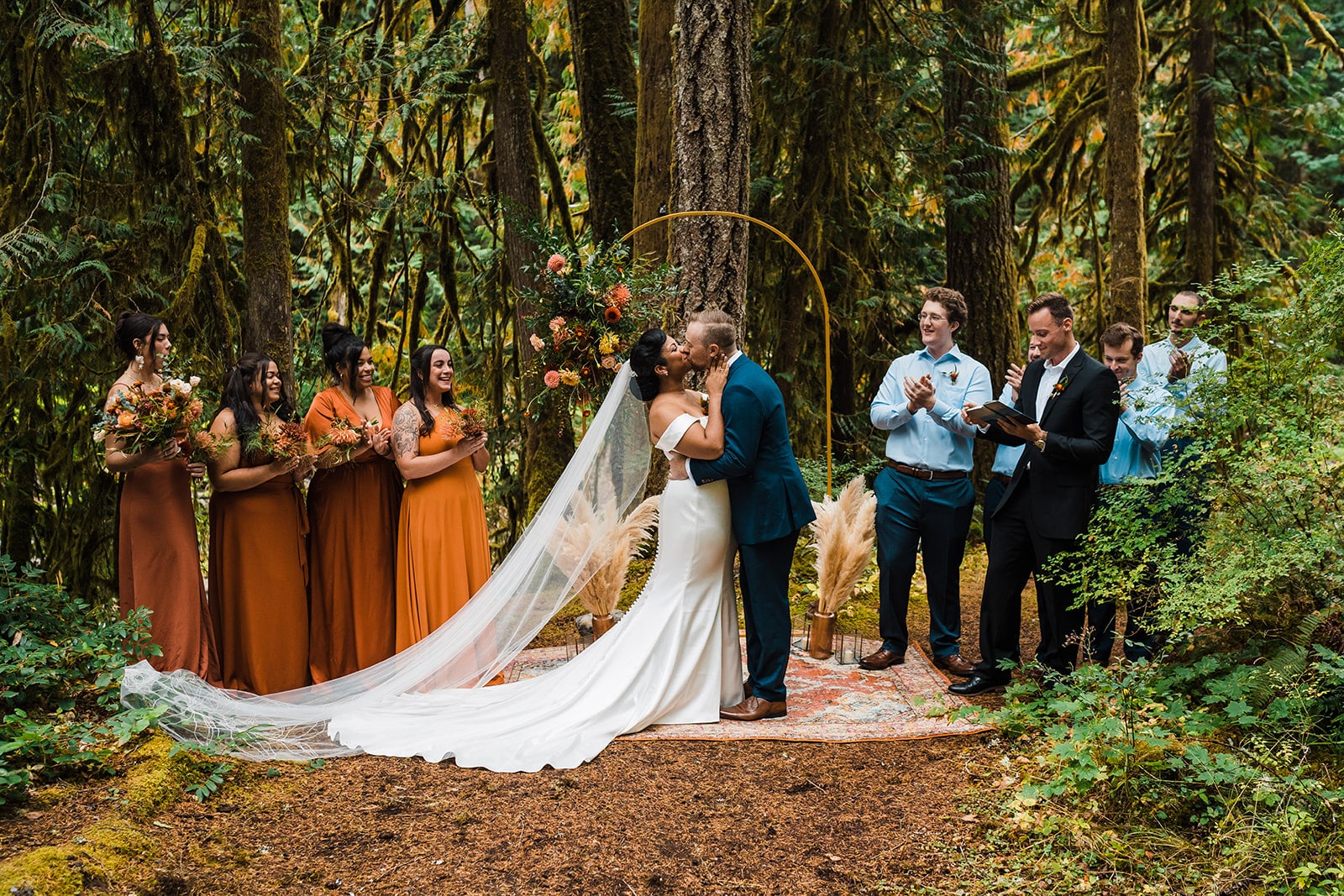 Bride and groom stand on a carpet in the forest and kiss while their wedding party cheers and claps. 