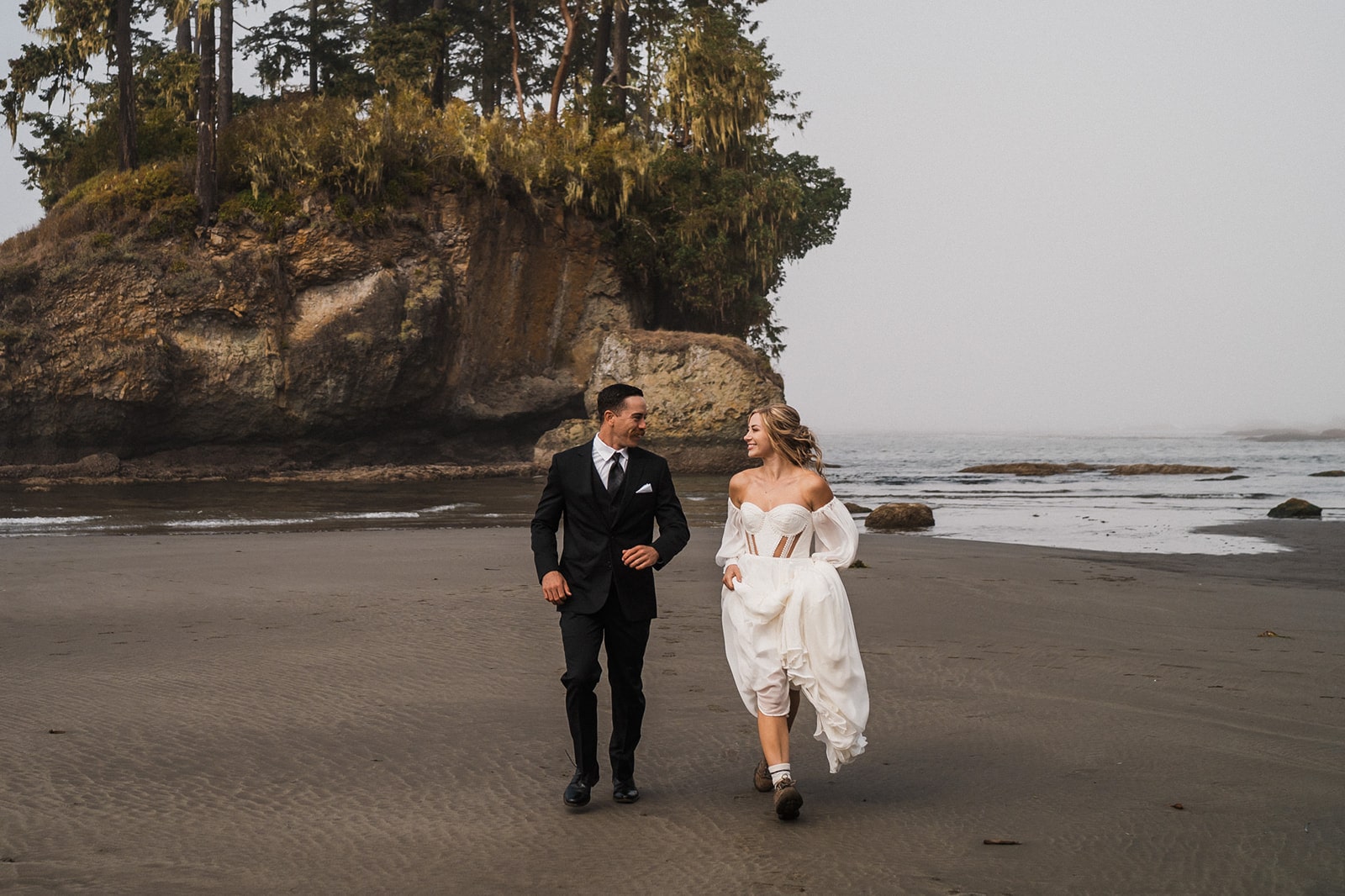 Bride lifts her elopement dress as she runs along the beach with groom during their elopement on the Olympic Coast. 