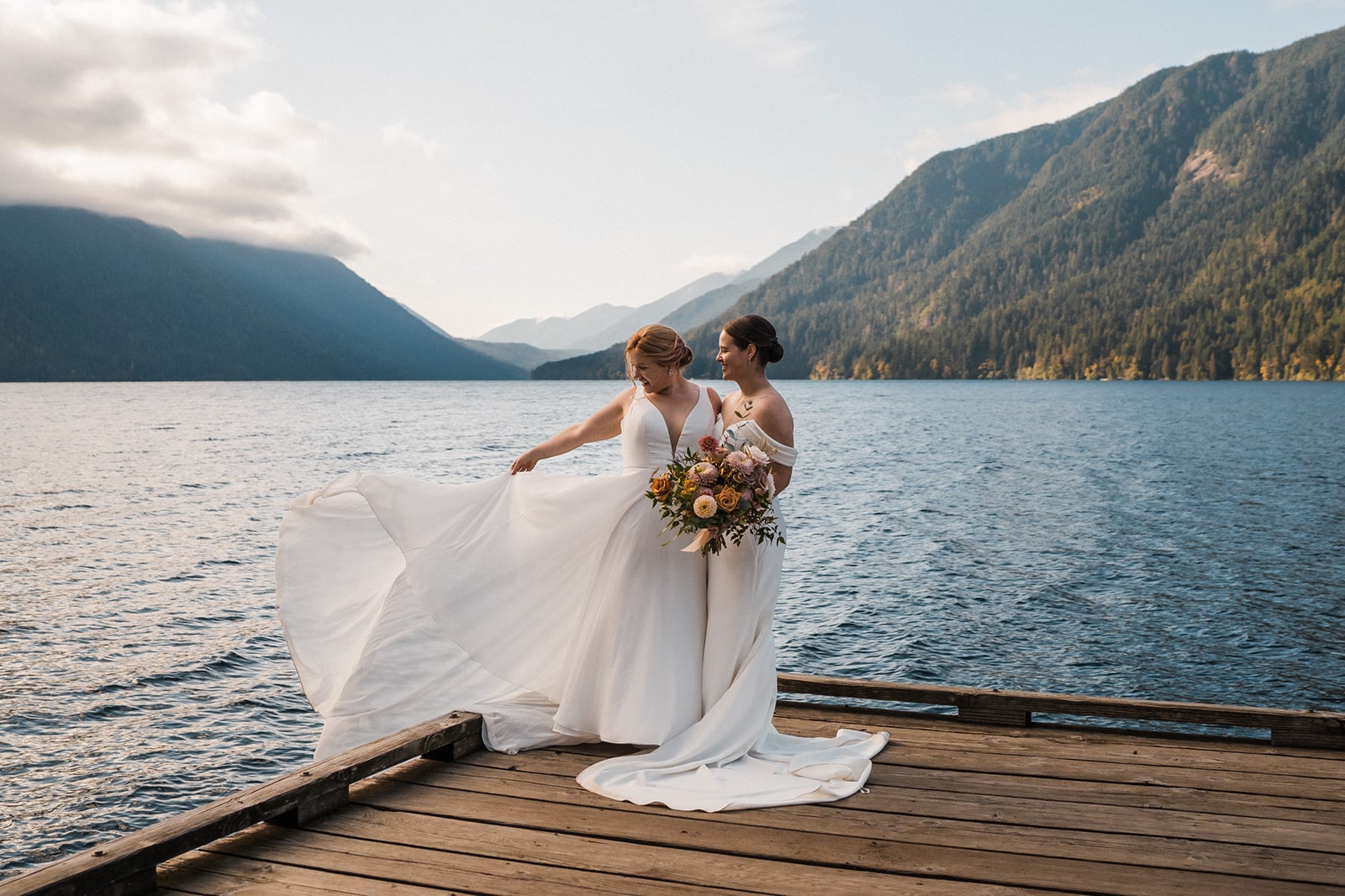 Brides stand on a dock in front of a lake while their white elopement dresses flow in the wind. 