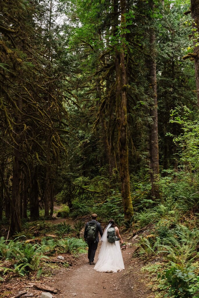 Bride and groom hold hands while wearing backpacks and hiking through the forest for their adventure wedding.