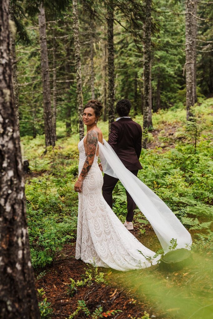 Brides hold hands as one partner wearing a white wedding dress turns to look back and her white cape veil flows in the wind. 