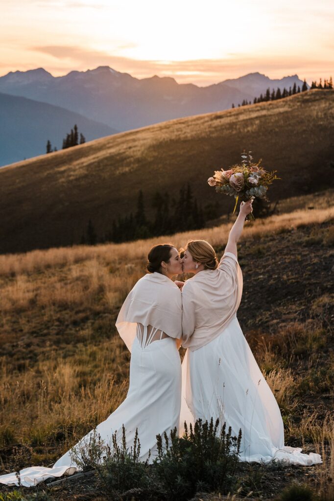 Two brides wearing white elopement dresses and shawls kiss while one holds up her elopement bouquet. 