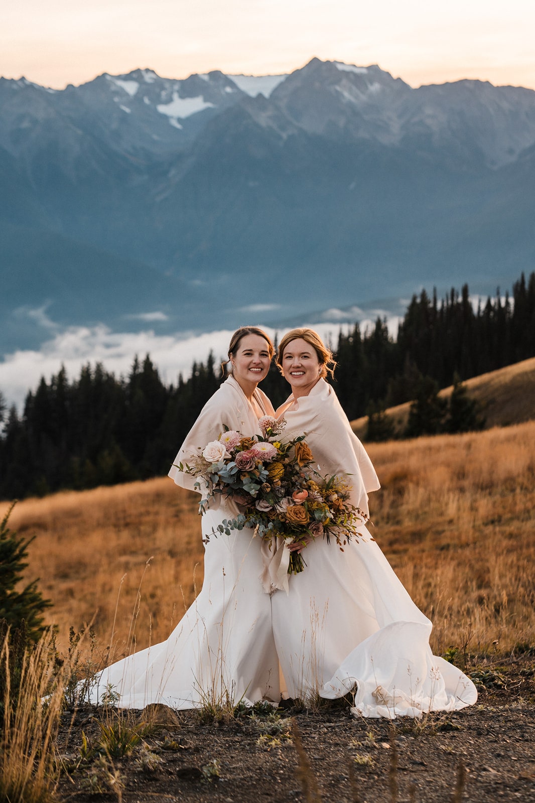 Two brides stand close together holding a large bouquet of flowers during their mountain elopement.