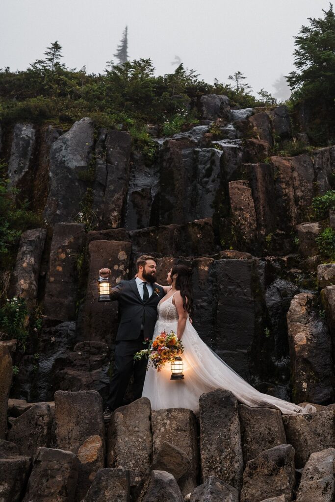 Bride and groom hold lanterns on a rocky mountainside trail during blue hour. 