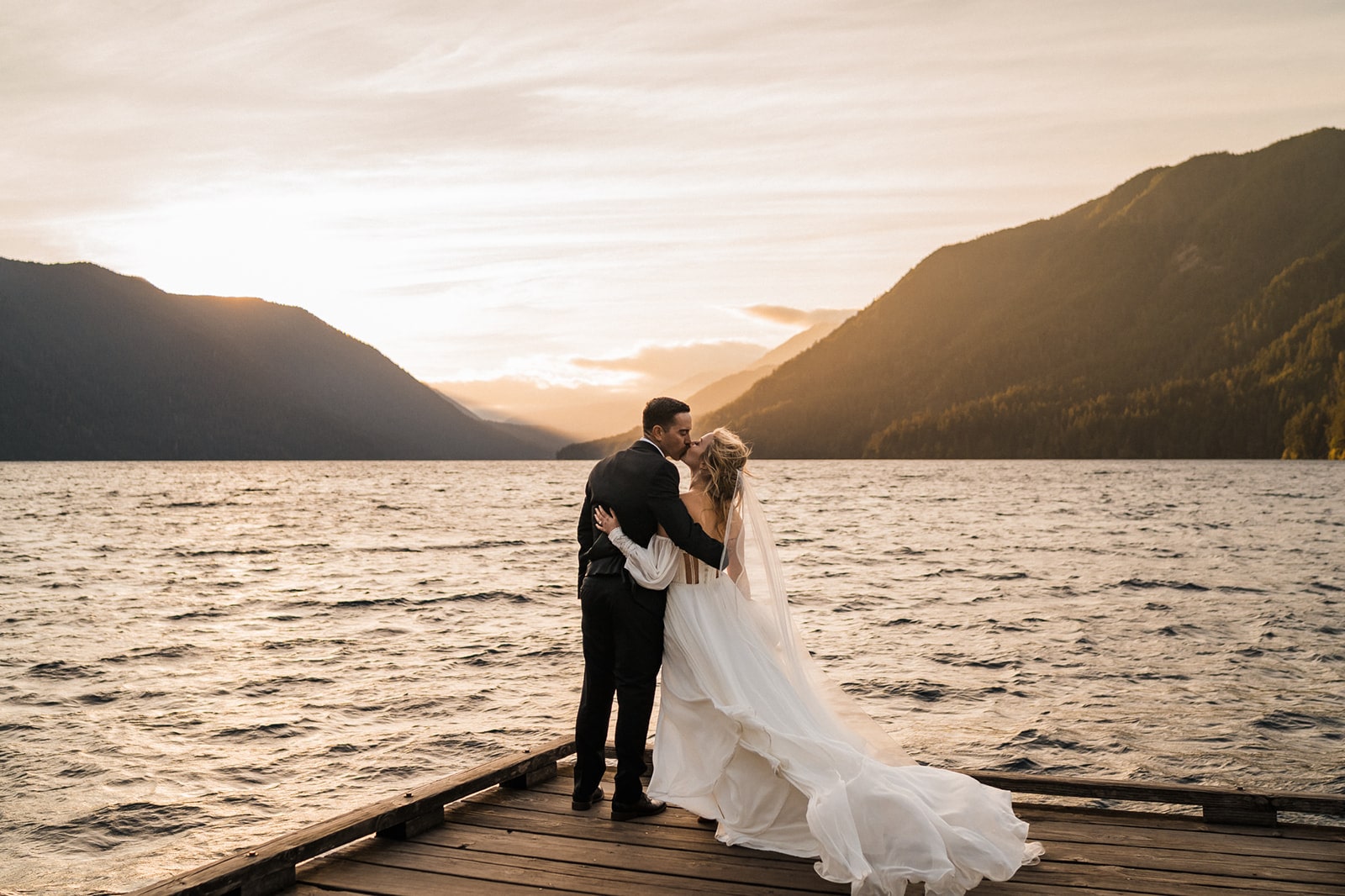 Bride and groom kiss on a dock overlooking a lake while bride's elopement dress flows in the wind. 