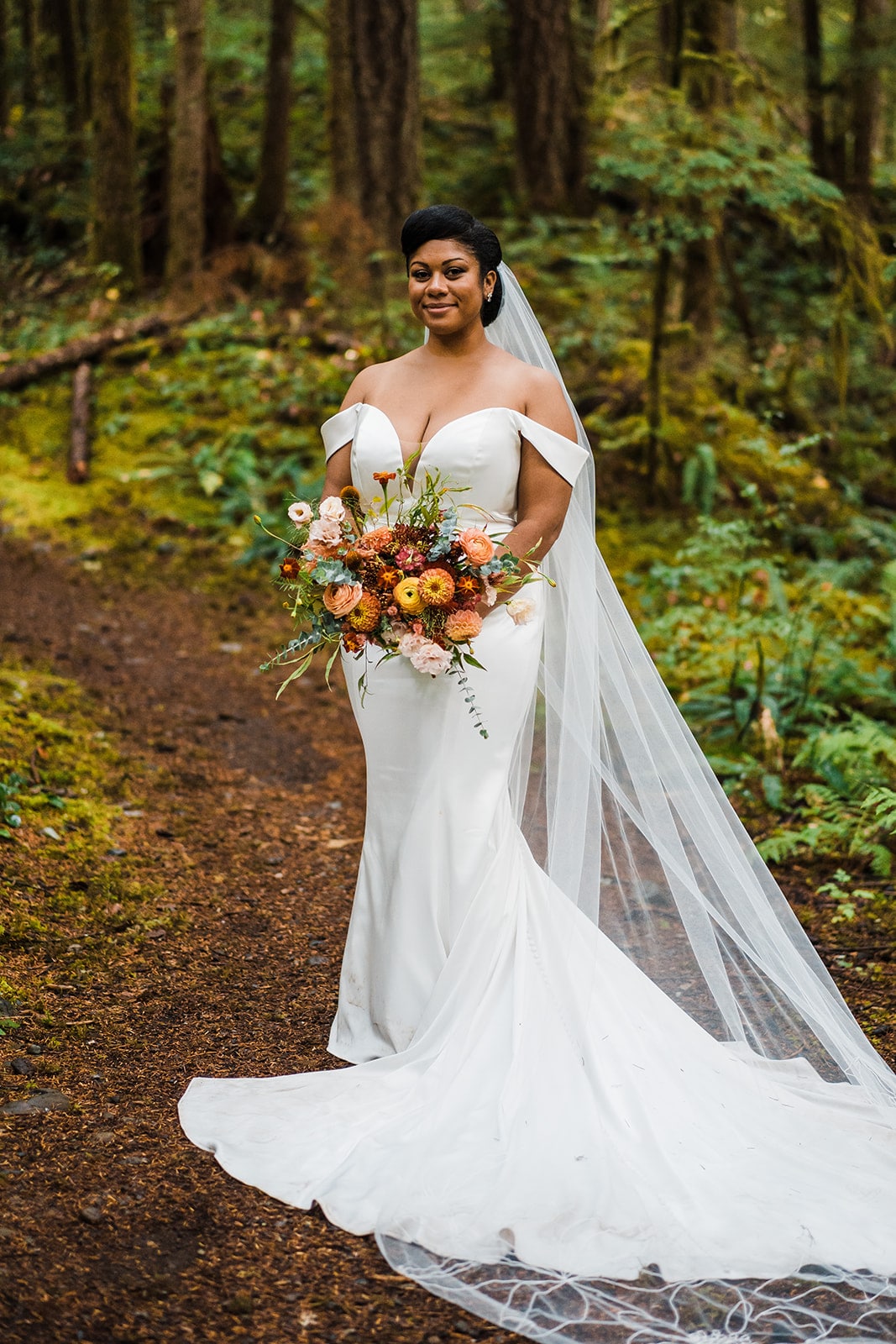 Bride stands on a forest trail holding a bouquet or orange and yellow wildflowers and her white wedding veil and dress train flow around her. 