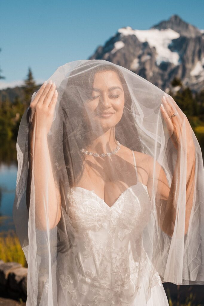 Bride plays with her tulle elopement veil as it catches the sunlight on a mountain trail. 