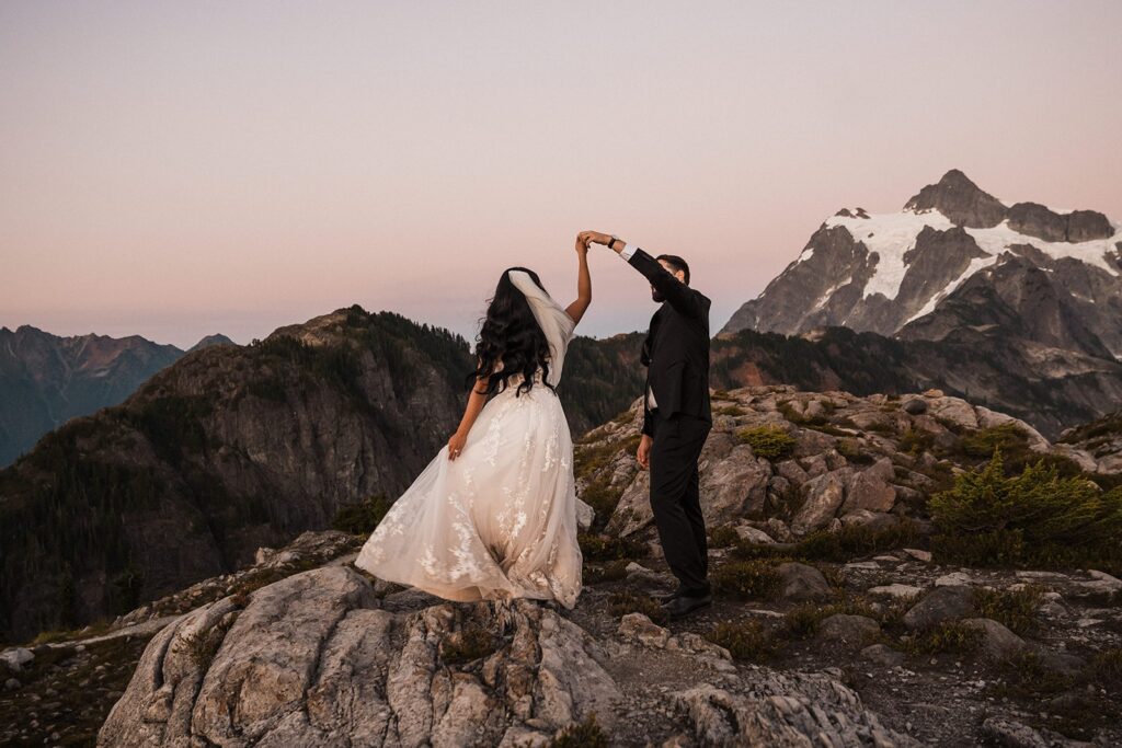 Groom twirls bride around while dancing on a mountain trail during their elopement in the North Cascades.