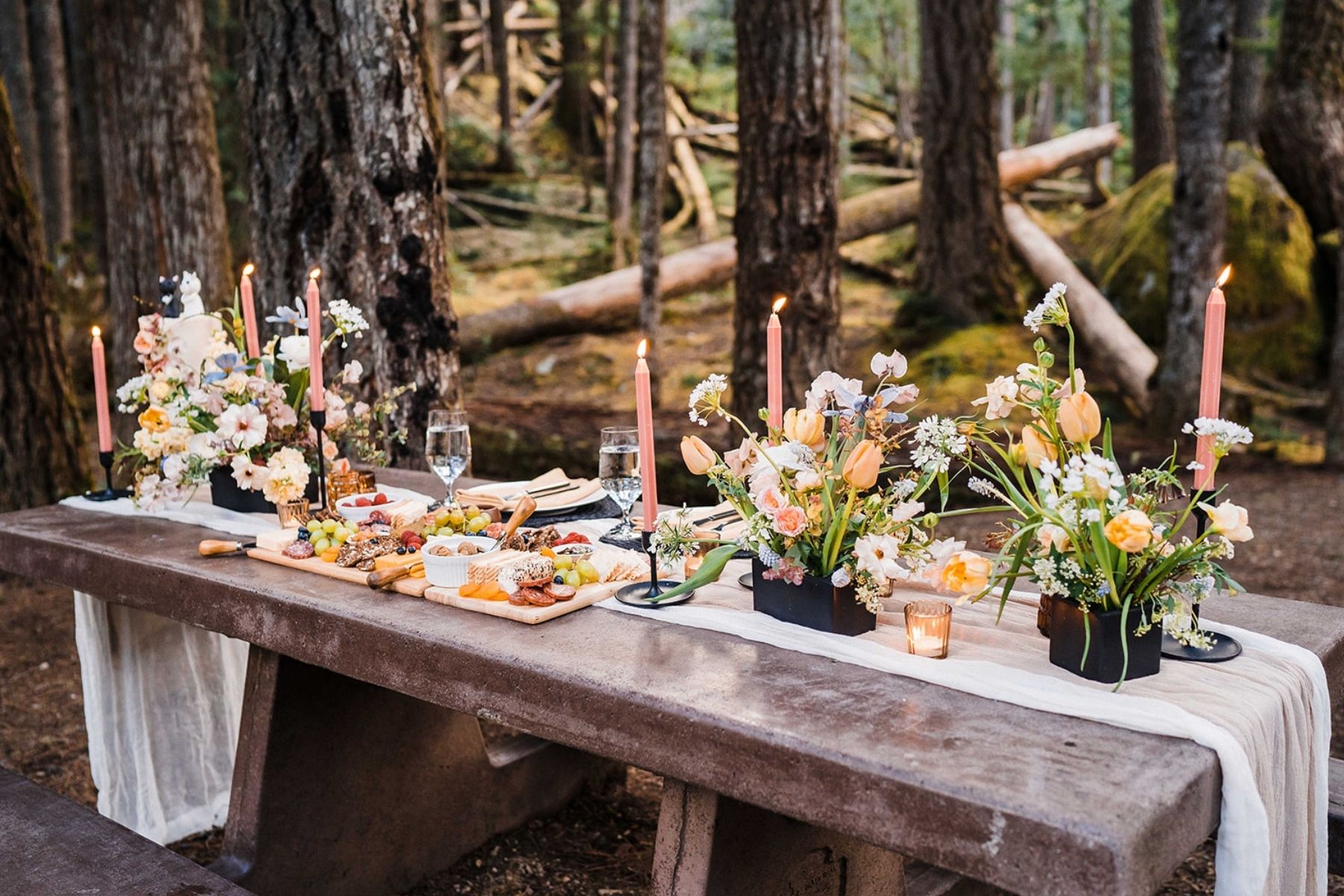 Pink and white flowers line a wood picnic table for an outdoor wedding reception dinner in the forest.