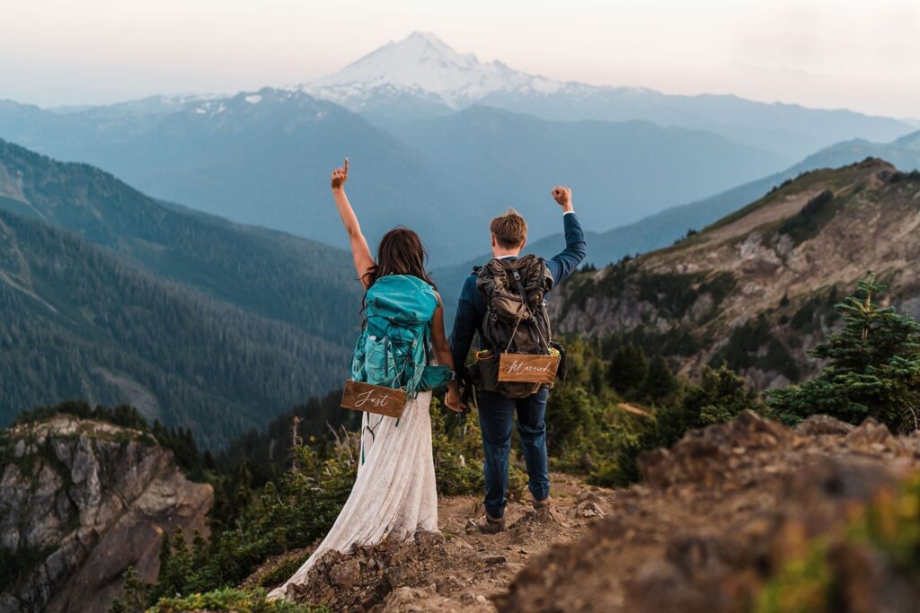 Bride and groom wear hiking backpacks with "Just Married" signs during their hiking elopement in the mountains
