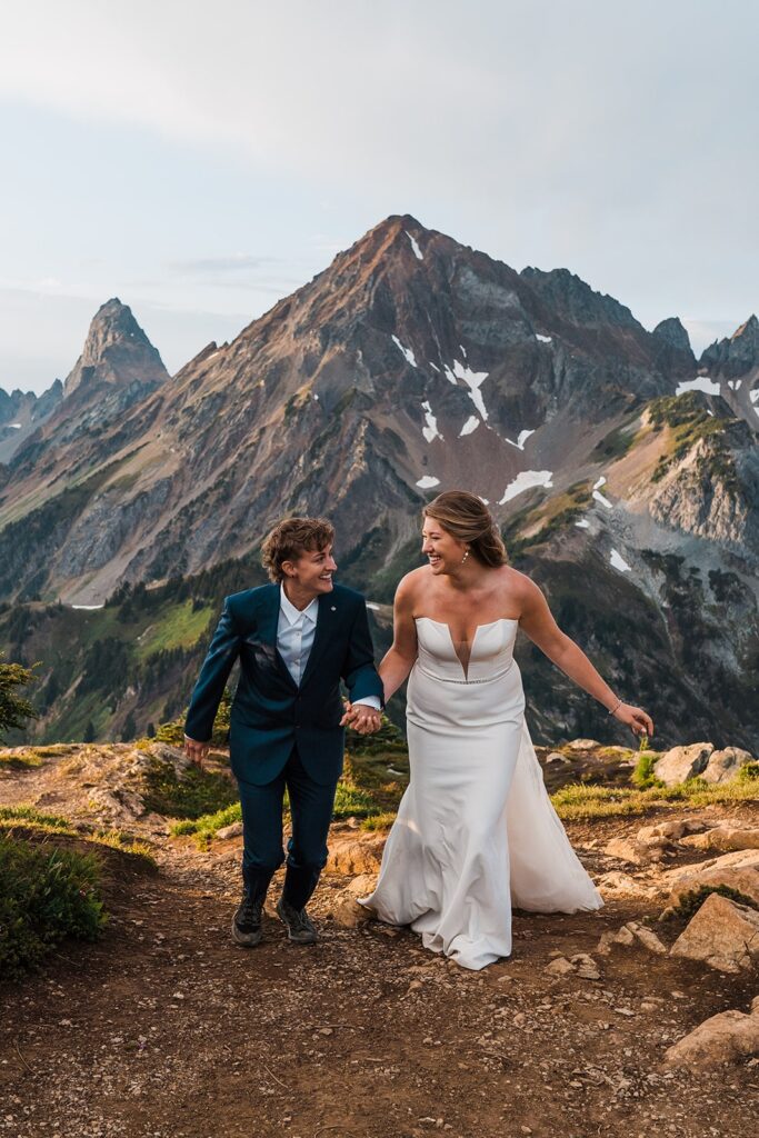 Brides hold hands and run across the mountain trail during their elopement in Washington