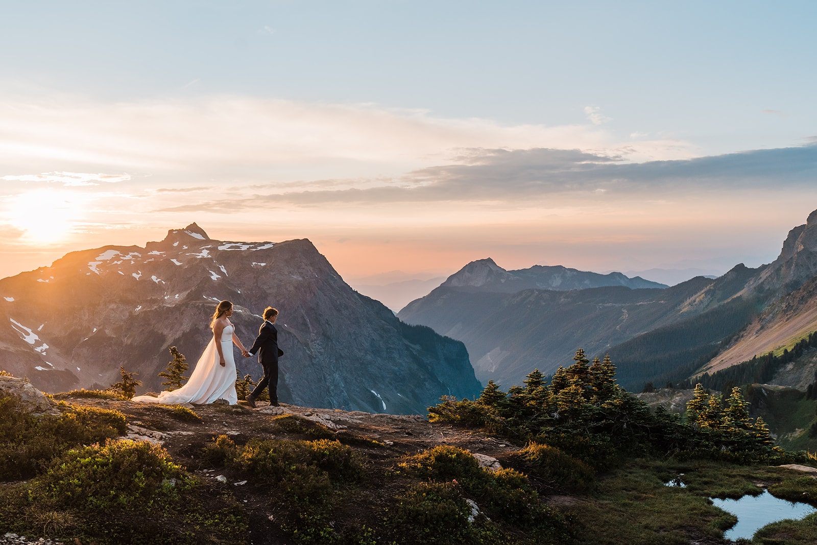 Two brides hold hands and walk across the mountain at sunset during their North Cascades elopement in Washington State.