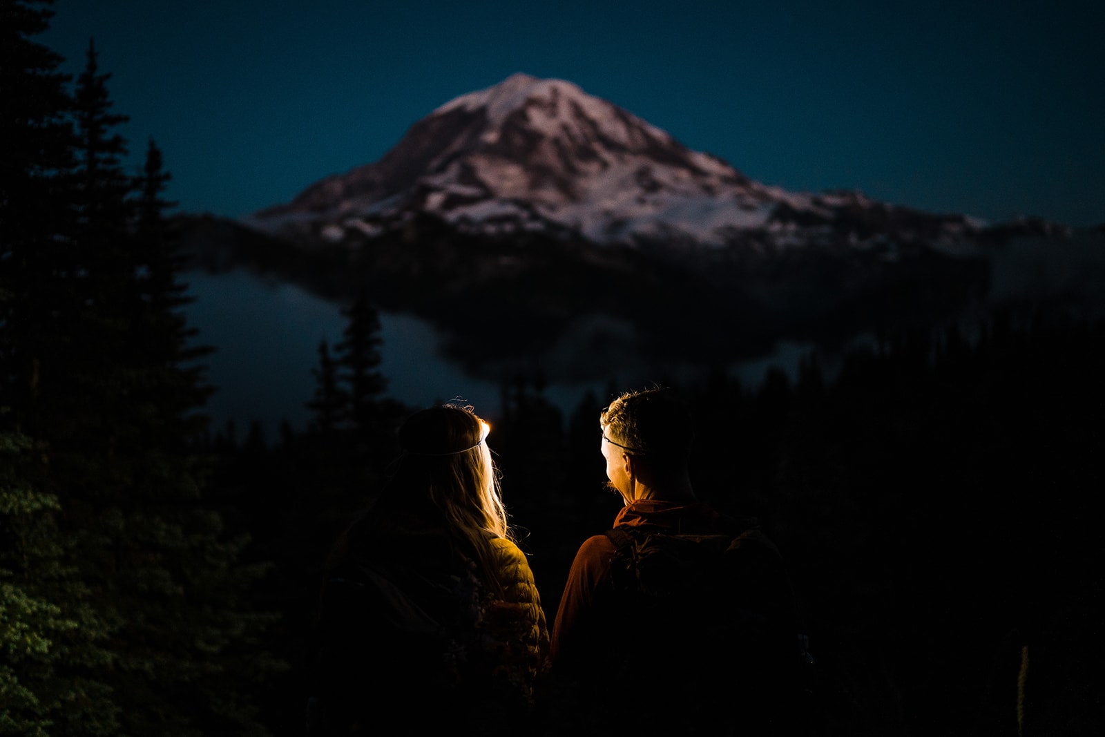 Bride and groom gaze off at Mount Rainier while wearing headlamps during blue hour.