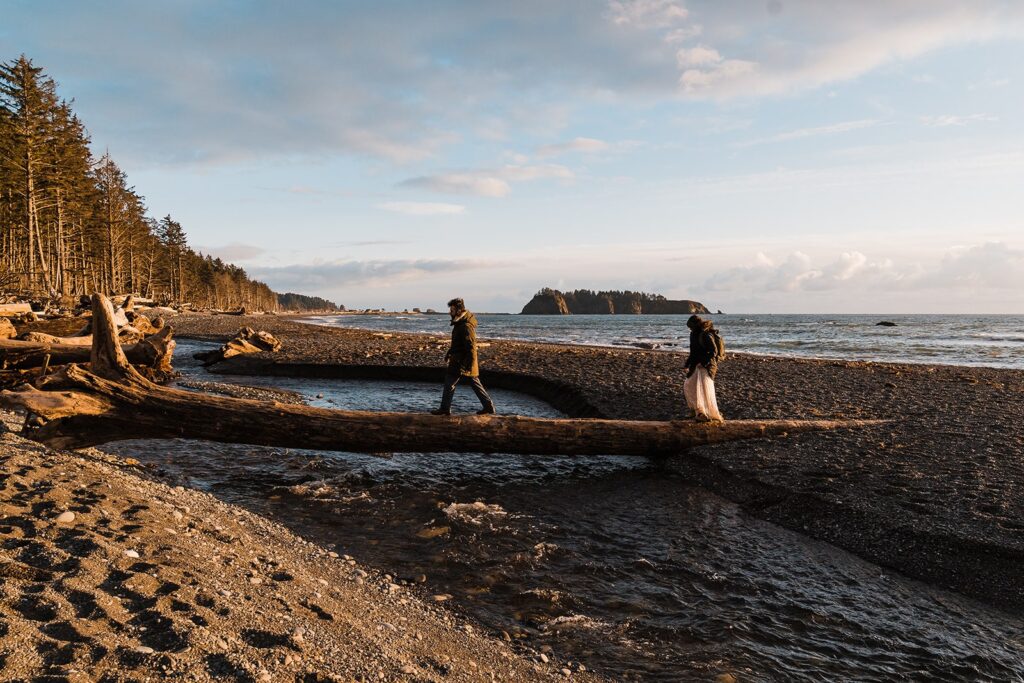 Bride and groom walk across a driftwood log at their beach elopement at Rialto Beach