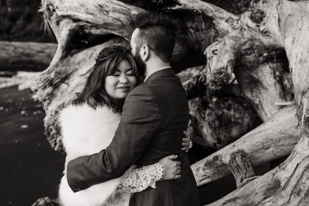Bride and groom hug in front of a pile of driftwood during their Washington spring elopement