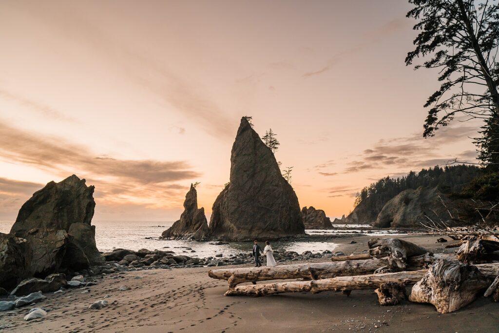 Bride and groom walk across a pile of driftwood during their sunset elopement photos on Rialto Beach