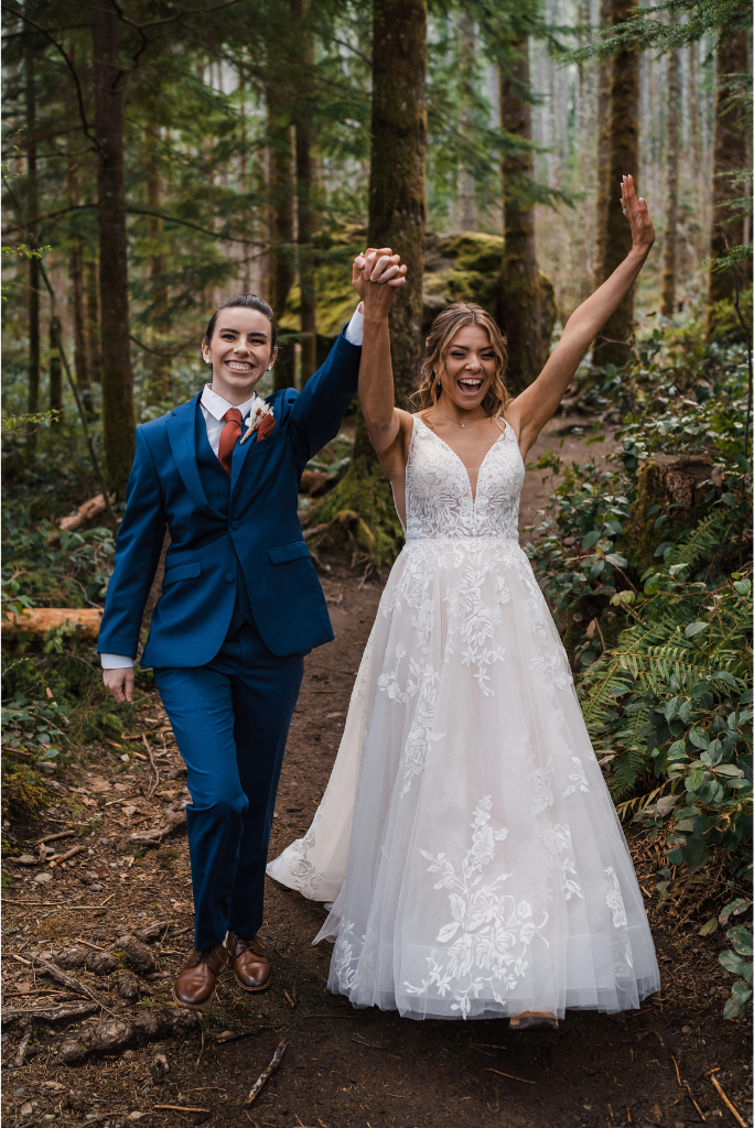 Brides raise hands and cheer while walking through the forest during their adventure wedding in Snoqualmie