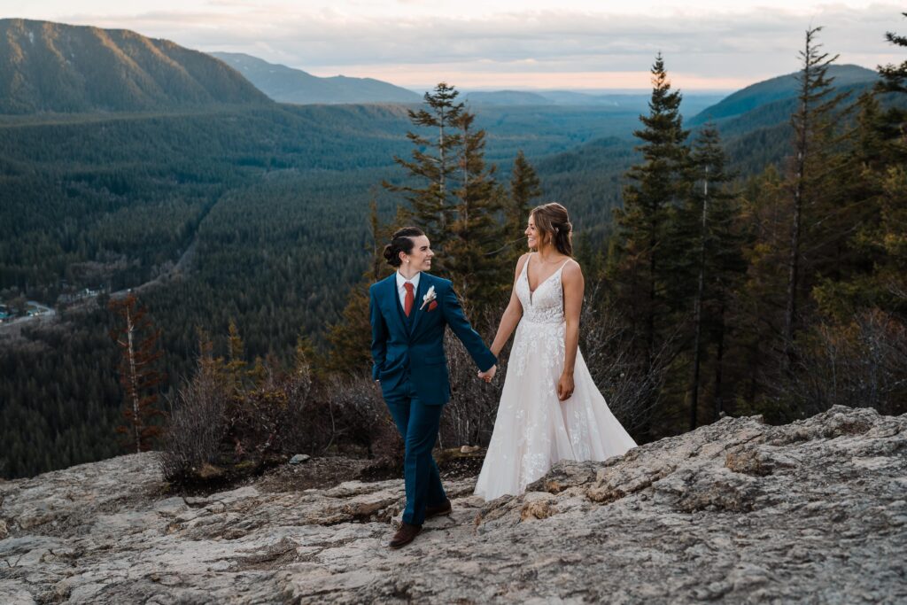 Brides hold hands while walking across a mountain during their adventure wedding in Snoqualmie