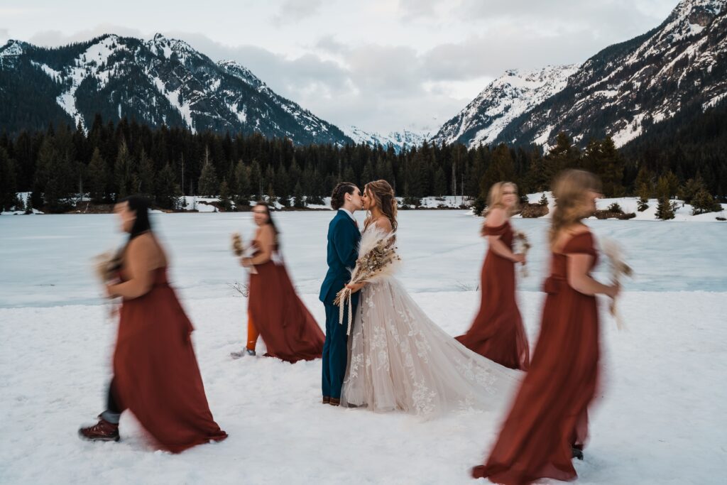 Brides kiss while bridesmaids walk around them in the snow