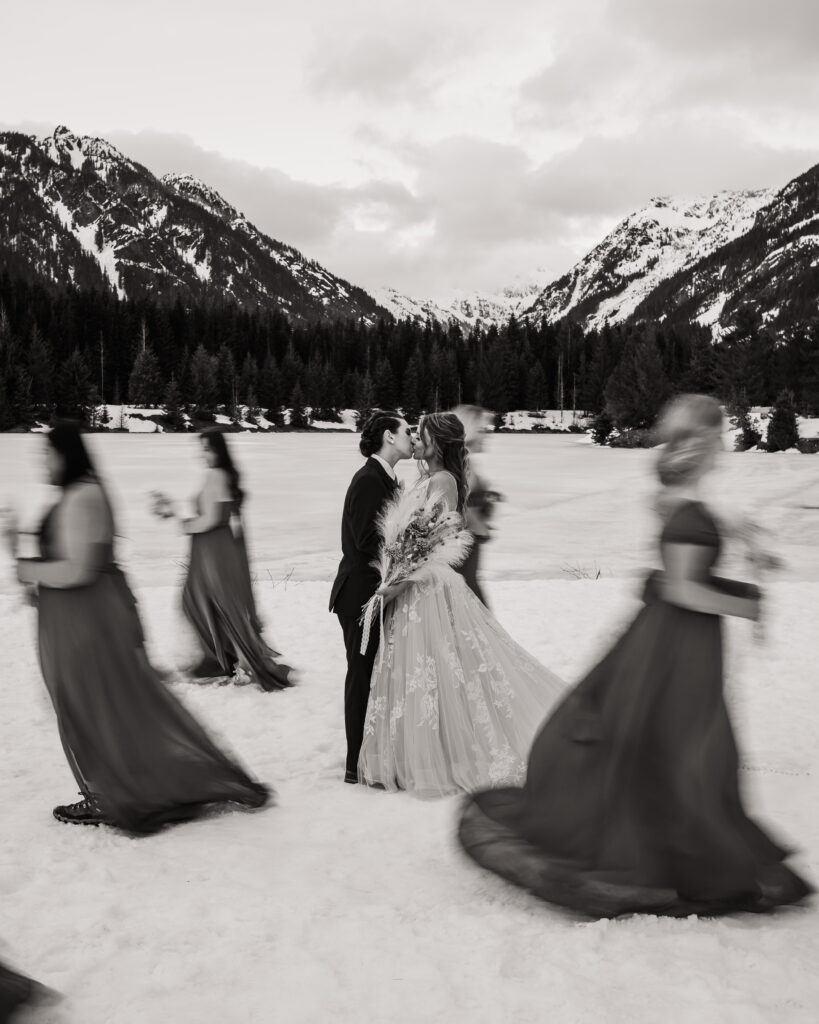 Brides kiss while bridesmaids walk around them in the snow