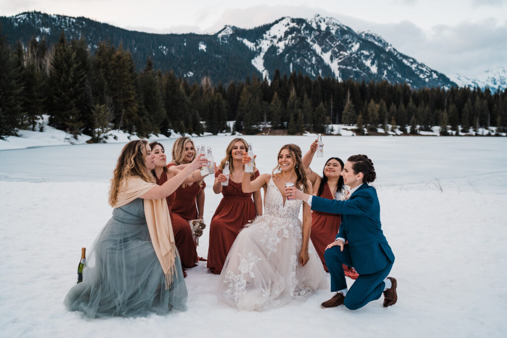 Brides and bridesmaids toast drinks to each other while kneeling in the snow