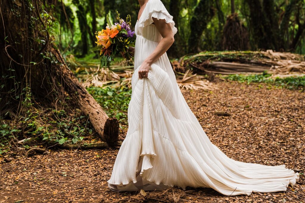 Bride holds train of white wedding dress while walking through the rainforest at her Hawaii elopement