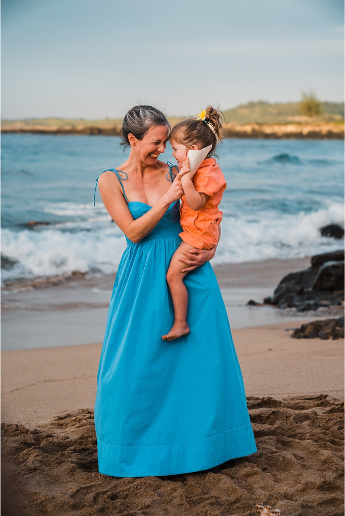 Elopement guest wearing blue sun dress and holding child in orange jumpsuit on the beach in Hawaii