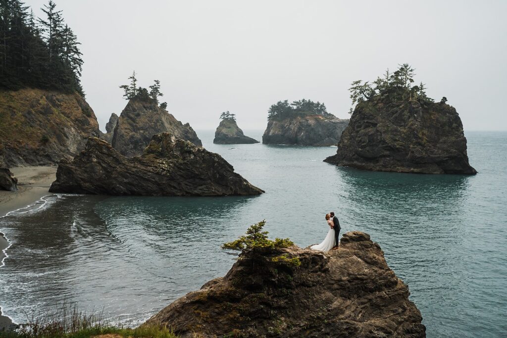Bride and groom stand on a rocky cliff and kiss during their beach elopement photos