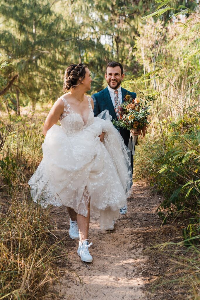 Bride lifts dress while groom holds flowers and they walk through a trail to their Kauai wedding ceremony location