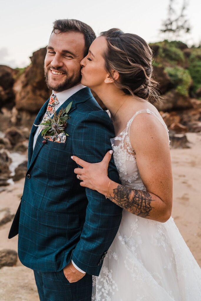 Bride kisses groom on the cheek during sunset photos in Kauai