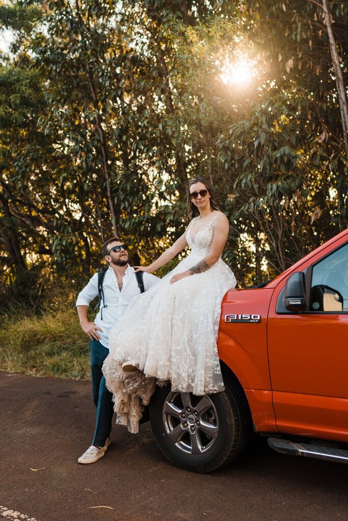 Bride sits on the hood of a Ford F150 while groom looks up at her during Kauai wedding photos
