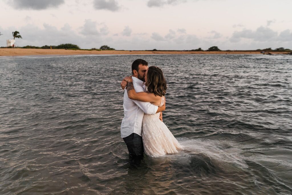 Bride and groom stand in the ocean and kiss during their sunset photos at their two-day wedding in Kauai