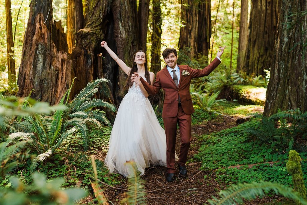 Bride and groom cheer as they walk through a forest trail during their redwoods elopement photos on the Oregon Coast