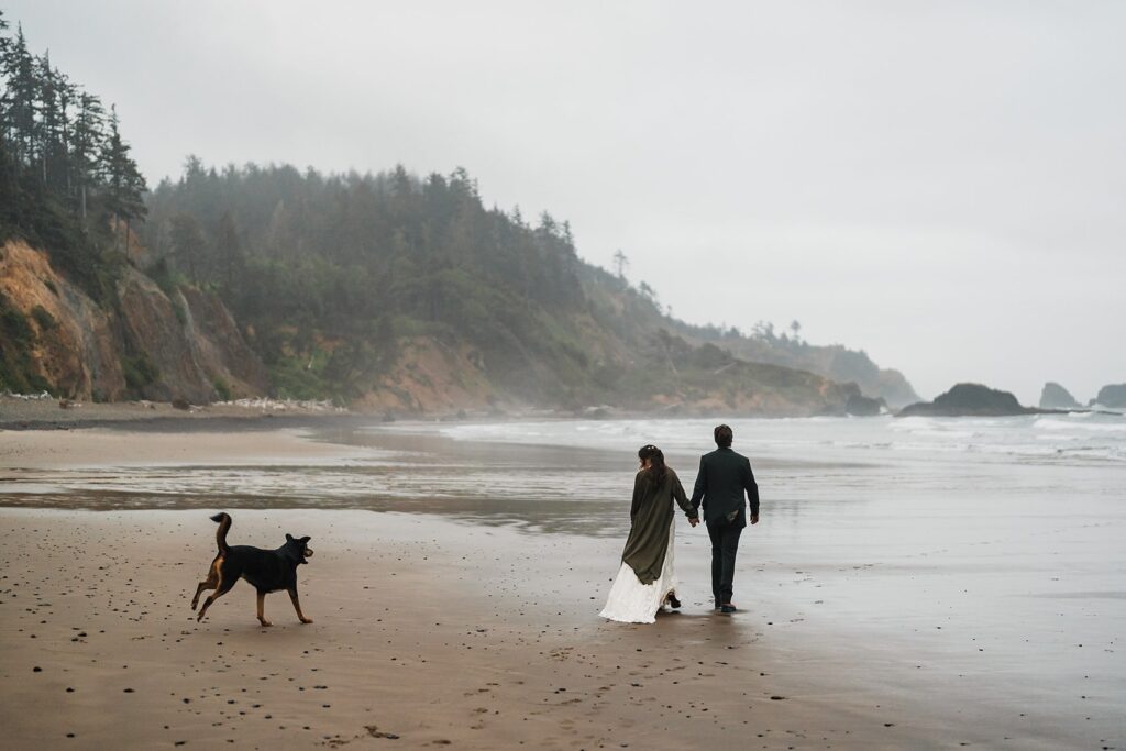 Bride and groom hold hands while walking across the beach at their Oregon Coast elopement
