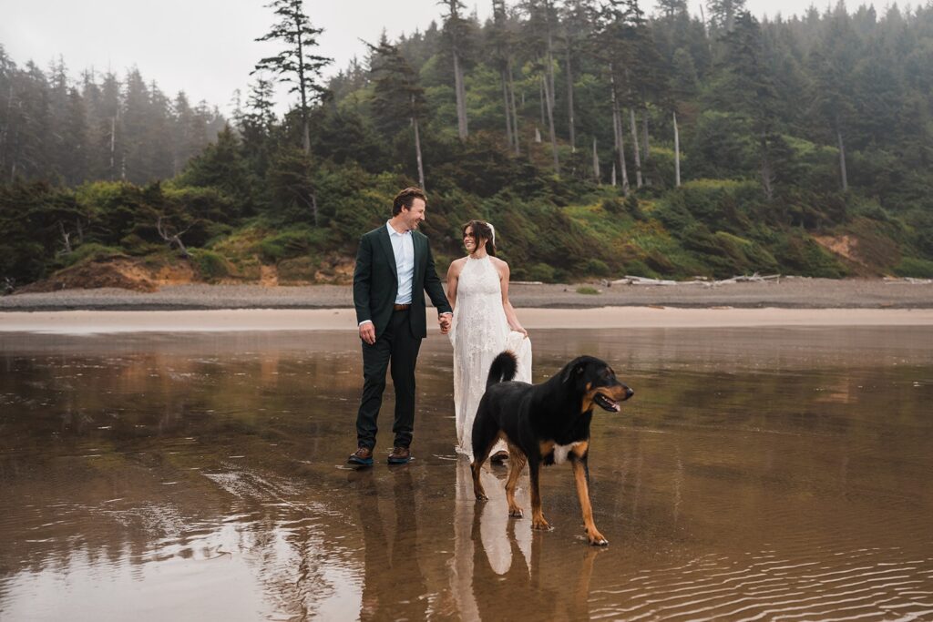 Bride and groom hold hands while walking across the beach with their dog during their Oregon Coast elopement
