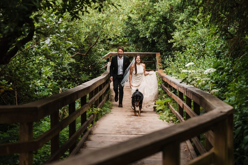 Bride and groom walk along a wooden bridge with their dog during their sunrise elopement photos on the Oregon Coast 