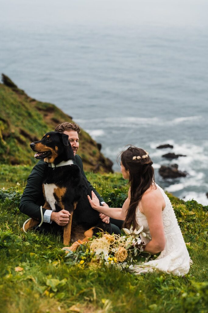 Bride and groom play with their dog while sitting on a cliff overlooking the ocean during their elopement on the Oregon Coast 