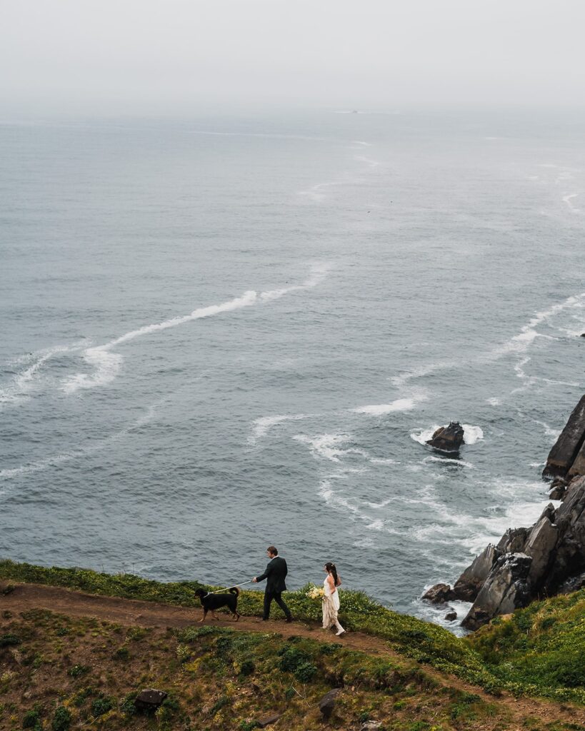 Bride and groom walk with their dog on an oceanside cliff trail during their Oregon Coast elopement 