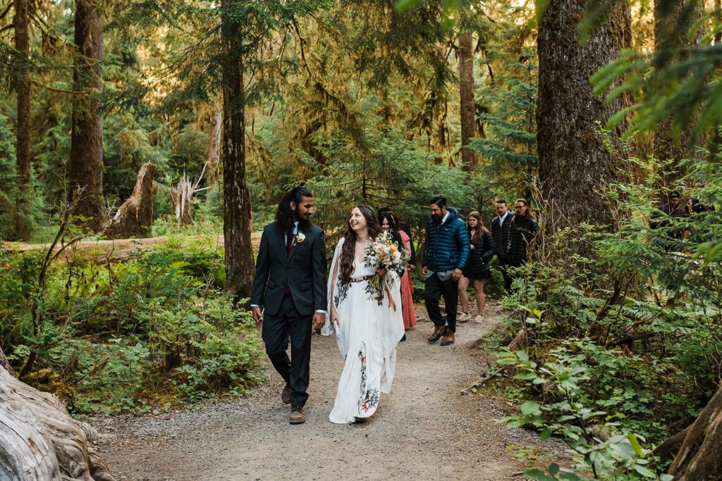 Bride and groom hold hands while walking through the forest with their wedding guests