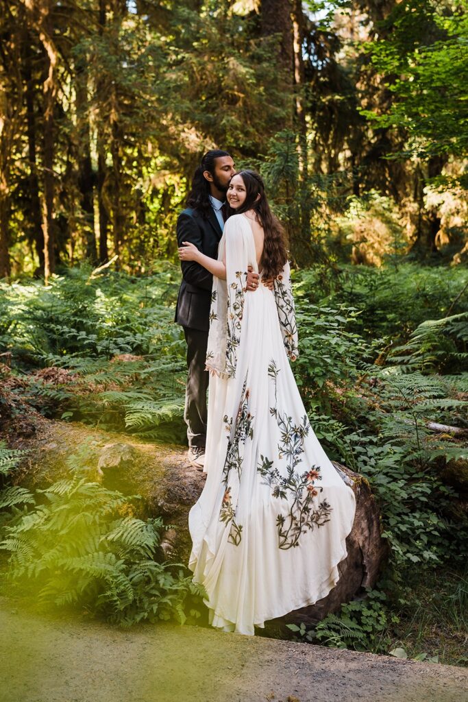 Bride and groom stand on a fallen tree with bride's cascading wedding dress train with wildflower embroidery flowing over the edge