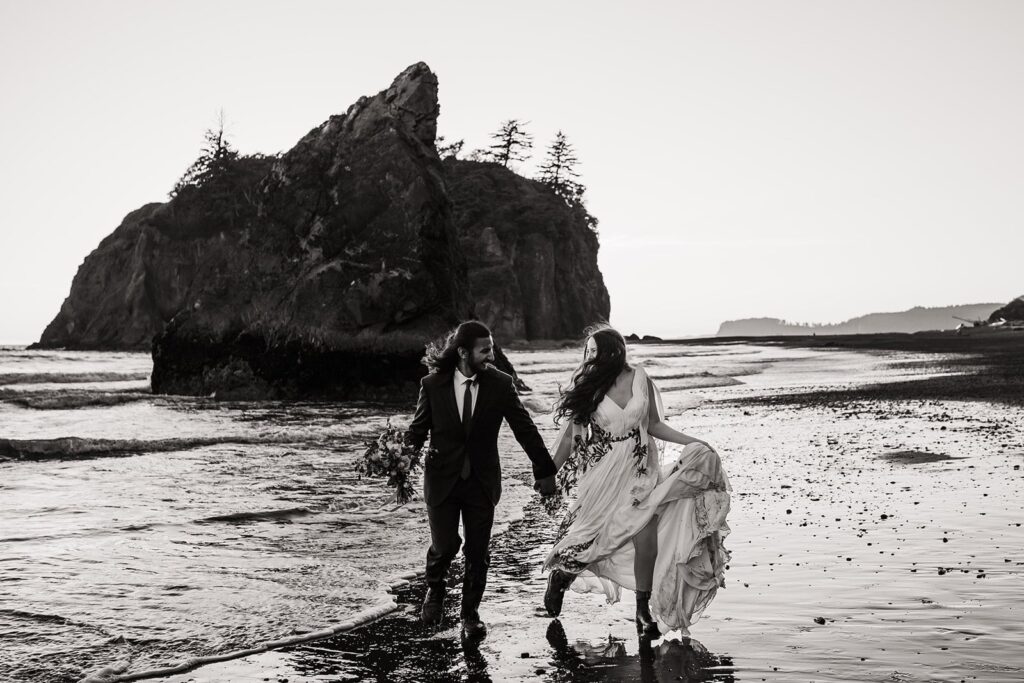 Bride and groom hold hands while running through the waves at Ruby Beach