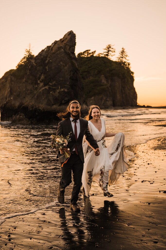 Bride and groom hold hands while running through the waves at Ruby Beach