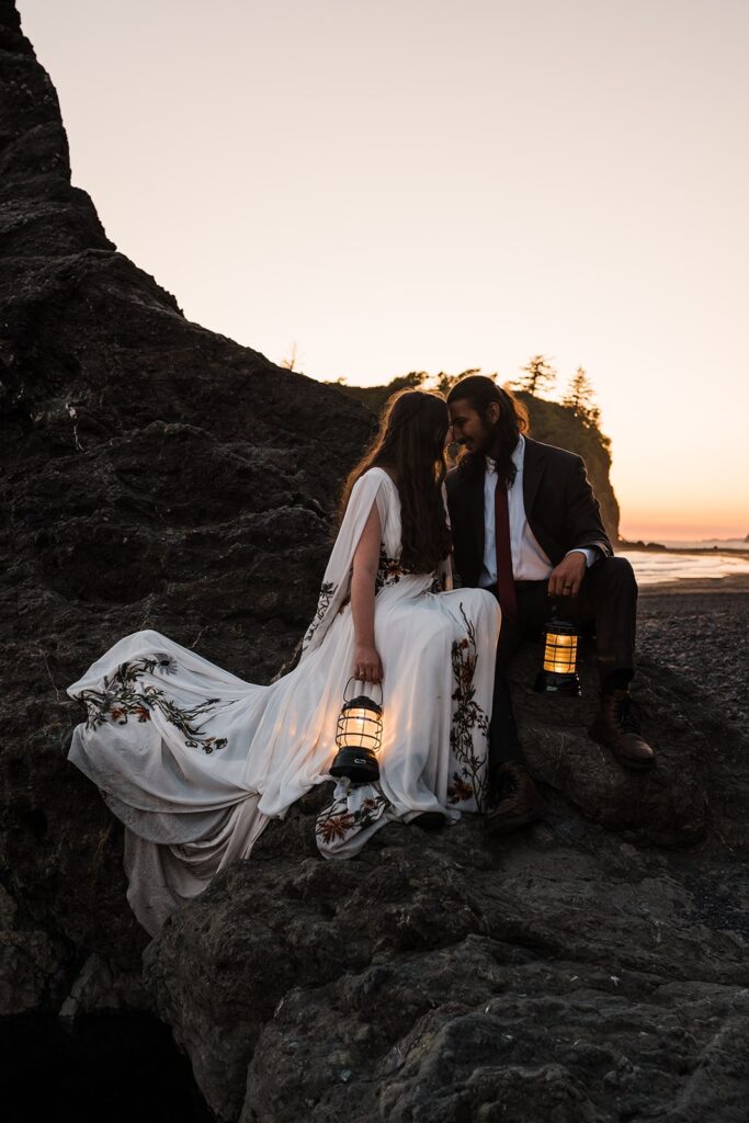 Bride and groom sit forehead to forehead while holding lanterns at sunset on the beach