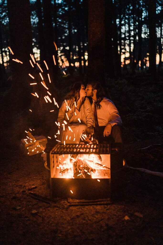 Bride and groom kiss while sitting in front of the fire roasting s'mores at their Hoh Rainforest wedding