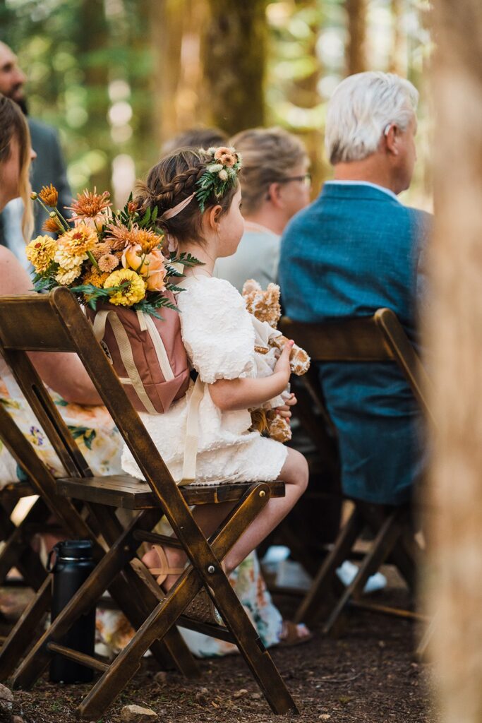 Flower girl wearing flower crown and flower backpack sitting in a wood chair during Olympic National Park wedding ceremony