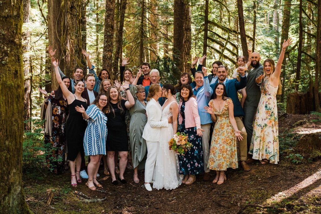 Brides kiss while friends and family cheer during family photos in the forest