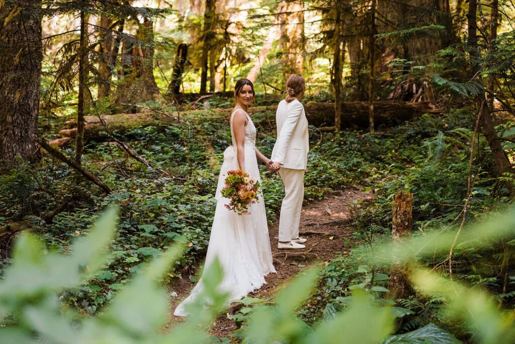 Brides hold hands while walking through the forest during their Olympic National Park wedding