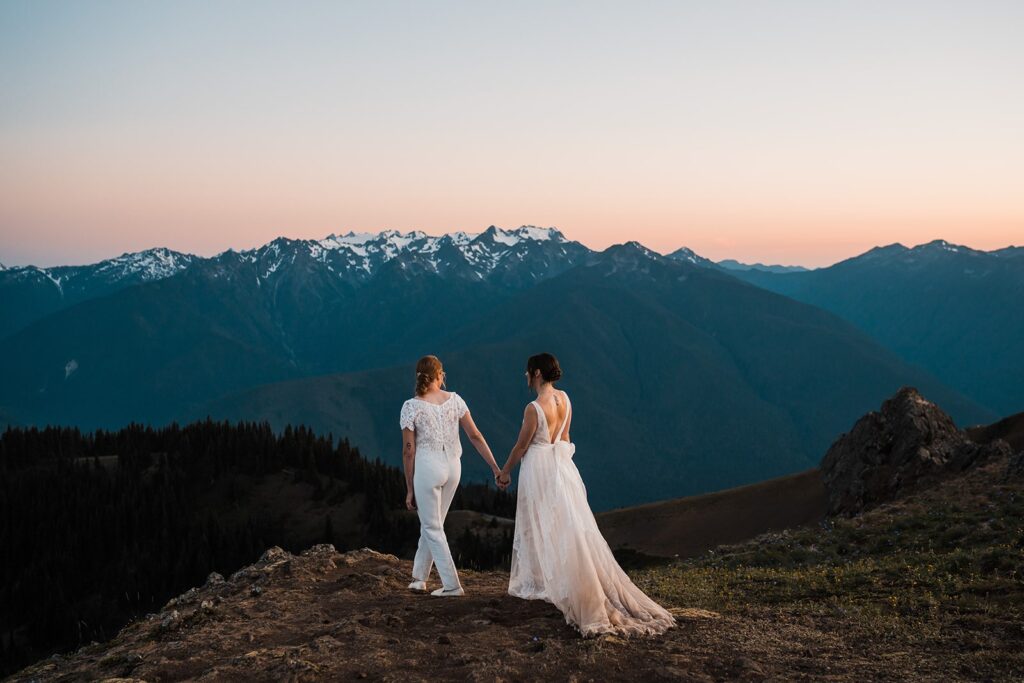Brides hold hands while walking across the field during their wedding photos in Olympic National Park